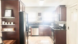 Une cuisine avec des meubles en bois foncé, un réfrigérateur en acier inoxydable, une cuisinière à gaz et un four à micro-ondes. On y trouve des étagères blanches où sont rangés des ustensiles de cuisine, ainsi qu’une fenêtre avec des stores.