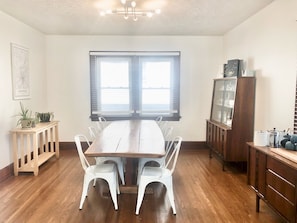 Une salle à manger avec une table en bois et des chaises blanches, un buffet en bois avec des portes en verre et une fenêtre avec des stores.