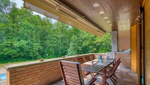 An outdoor patio with a brick wall, wooden chairs, a table, and a view of a forest. - Villa Bella Elena Private Swimming Pool (Vergiate)