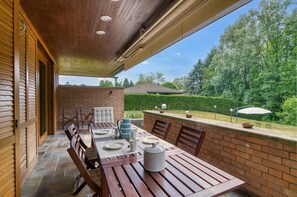 Eine Terrasse im Freien mit Backsteinmauer, Holztisch und Stühlen, mit Blick auf einen Garten und ein Gebäude in der Ferne.
