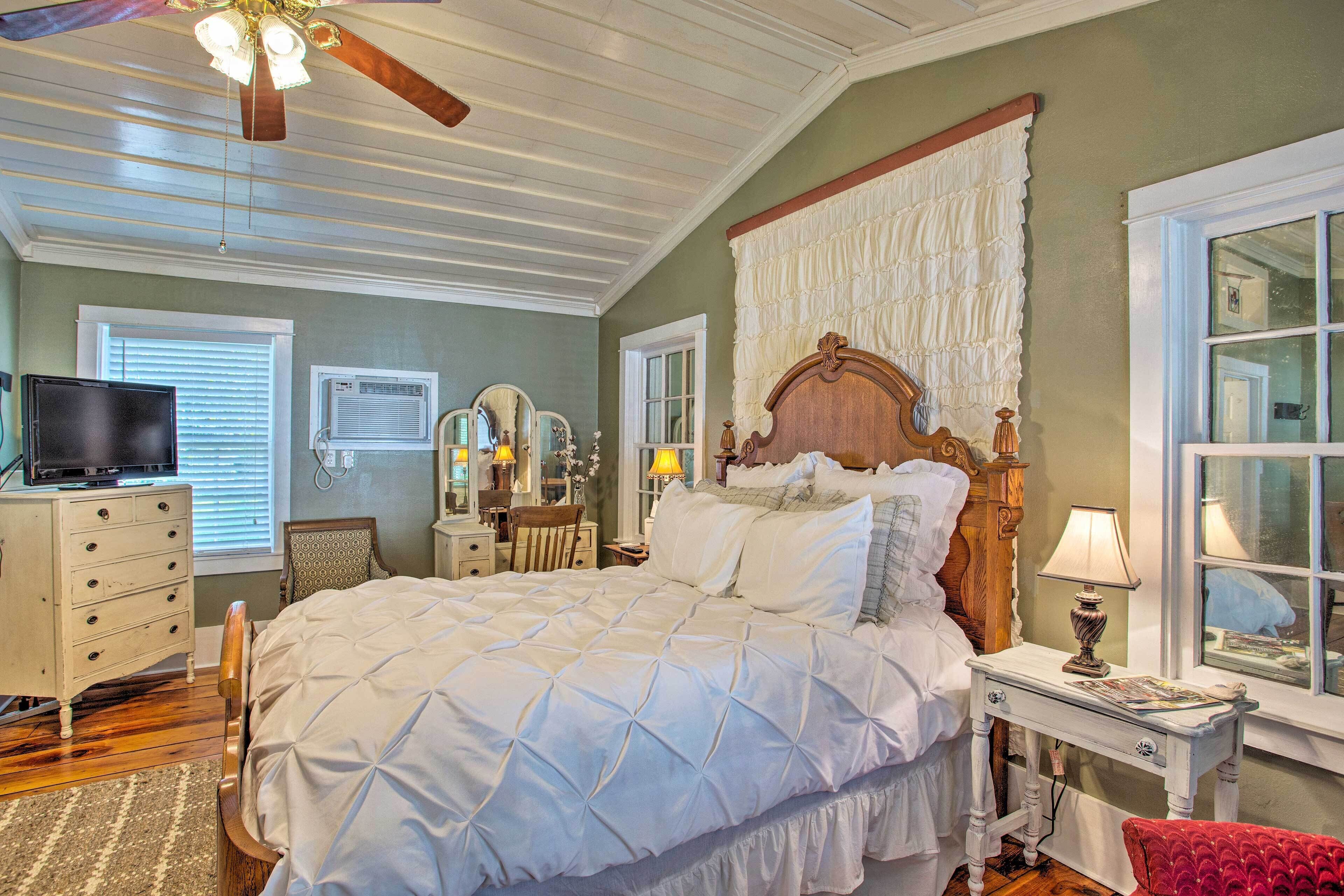 A bedroom with a large bed, a dresser, a vanity with mirrors, a ceiling fan, and a window with blinds.