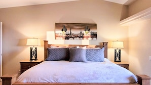 A bedroom with a wooden bed, two bedside tables with lamps, and a desert-themed painting above the bed. - "Hacienda Hangout"~ 5⭐RETREAT ~ PETS OK 🐾  VIEWS - POOL - SPA - BORDERS BLM  (Lake Havasu City)