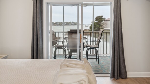 A bedroom with a large bed, a view of a balcony with chairs, and a body of water in the background. - Riverfront condo, Screen porch - Short walk across bridge to beach, shops, and restaurants! (Folly Beach)