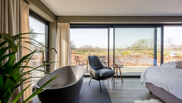 A modern hotel room with a large window, a black bathtub, a comfortable chair, a small table, and a neatly made bed. - Little Hayloft by Bloom stays -  charming barn with hot tub (Canterbury)