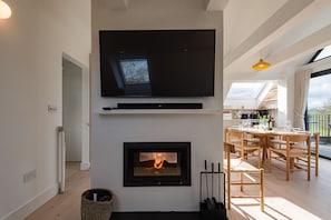 A modern living room with a flat-screen TV mounted above a fireplace, wooden dining table, and chairs.