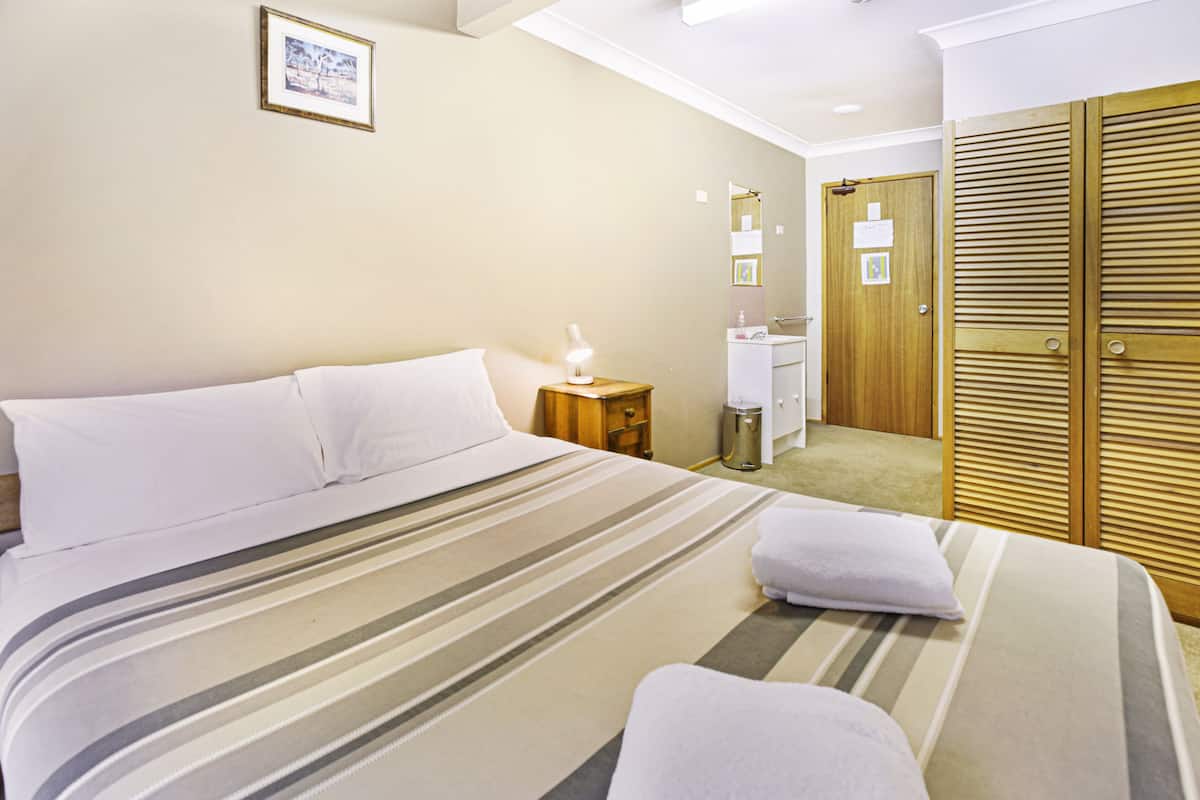A hotel room with a bed, a nightstand, a lamp, a bathroom with a sink and mirror, and a wooden wardrobe.