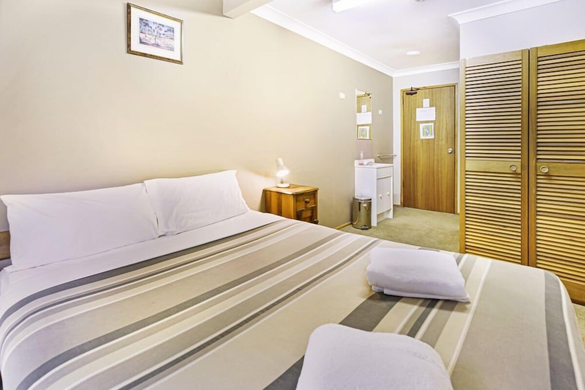 A hotel room with a bed, bedside table with a lamp, a wooden wardrobe, and a bathroom with a mirror and sink.