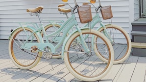 Een turquoise fiets met rieten manden en een bruine zitting geparkeerd op een houten terras voor een wit huis met een groene raamkozijn.