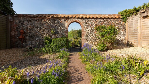 Ein steinerer Bogen führt in einen Garten mit Kiesweg und violetten Blumen.
