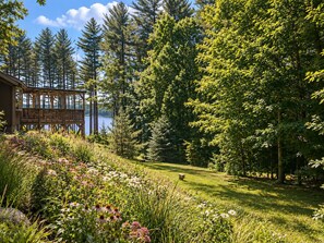 A wooden deck overlooking a lake with dense trees and a grassy area with wildflowers. - Lake View Berkshire Retreat – Private Setting Near Lenox (Lee)