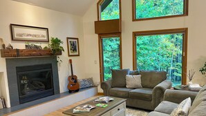 A living room with a fireplace, a couch, a coffee table, and large windows with a forest view. - Lake View Berkshires Retreat - Private & Minutes to Lenox (Lee)
