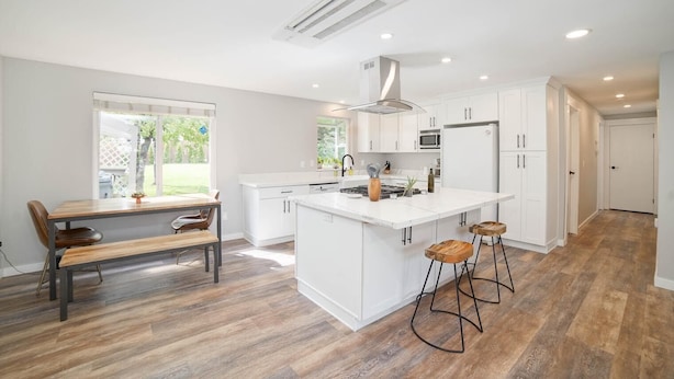 A modern kitchen with white cabinetry, a central island, and a dining area with a wooden table and chairs. - Beautiful Stylish Home With Hot Tub (Puyallup)