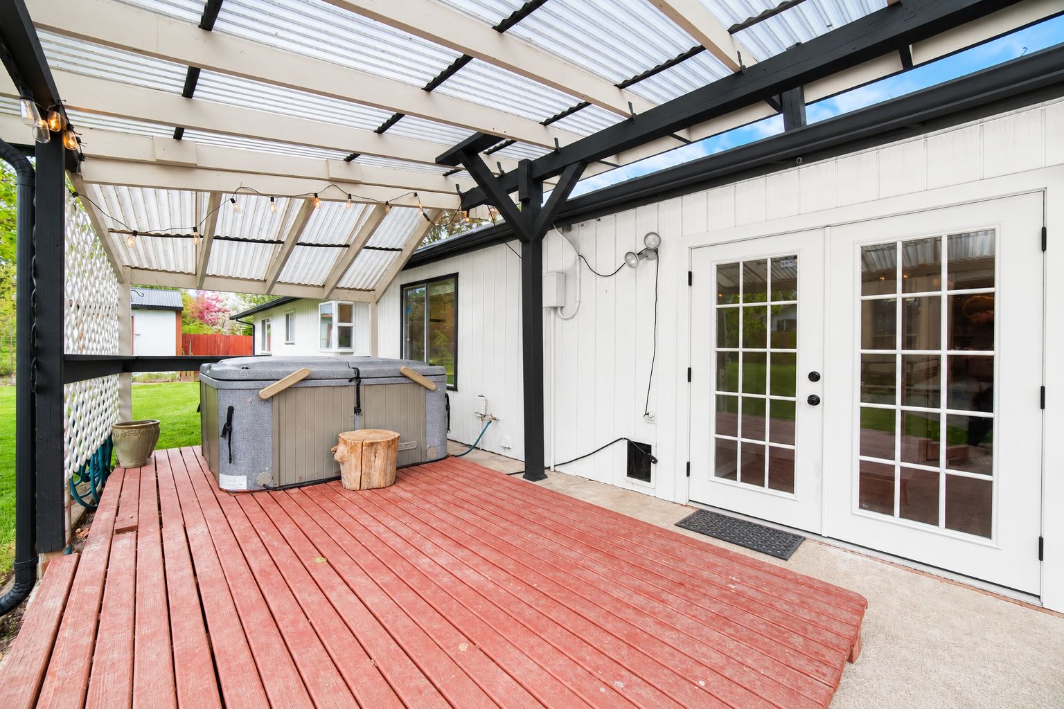 Een overdekt terras met een jacuzzi, houten terras en een wit gebouw met glazen deuren.