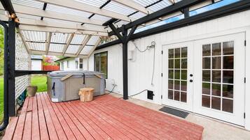 Een overdekt terras met een jacuzzi, houten terras en een wit gebouw met glazen deuren.