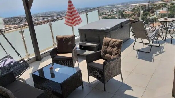 An outdoor patio area with wicker furniture, a glass table, and a view of a cityscape. - Panoramic Holidays - Deluxe 3 - Apartment with Outdoor Jacuzzi - Sea View (Peyia)