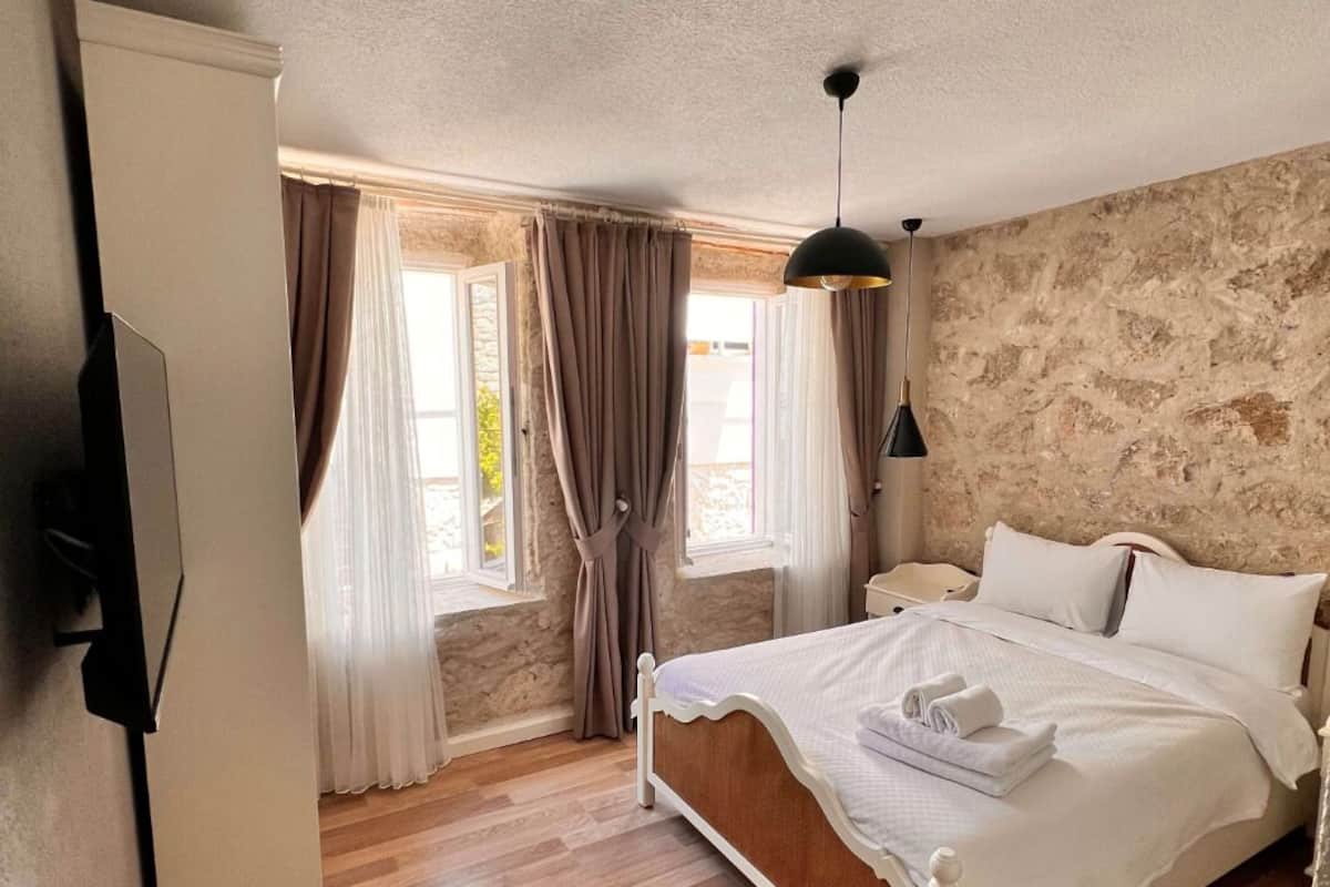 A bedroom with a wooden floor, a bed with white bedding, a black pendant light, and a stone wall.