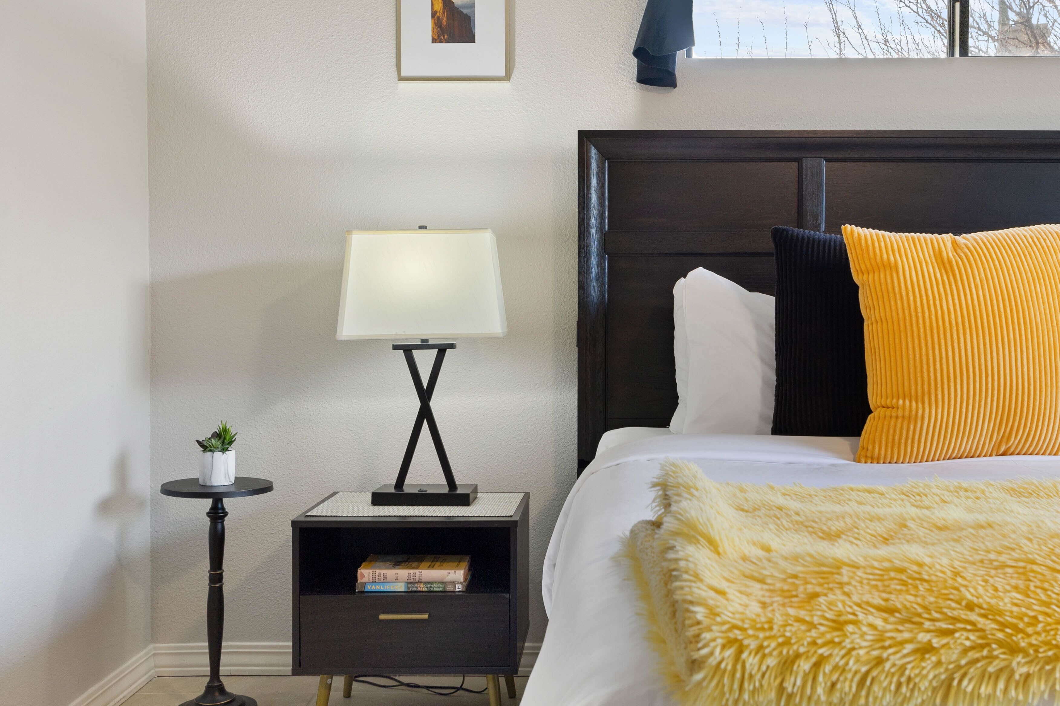 Un dormitori ben ordenat amb un llit, una taula de nit, una làmpada i una petita estant amb llibres.