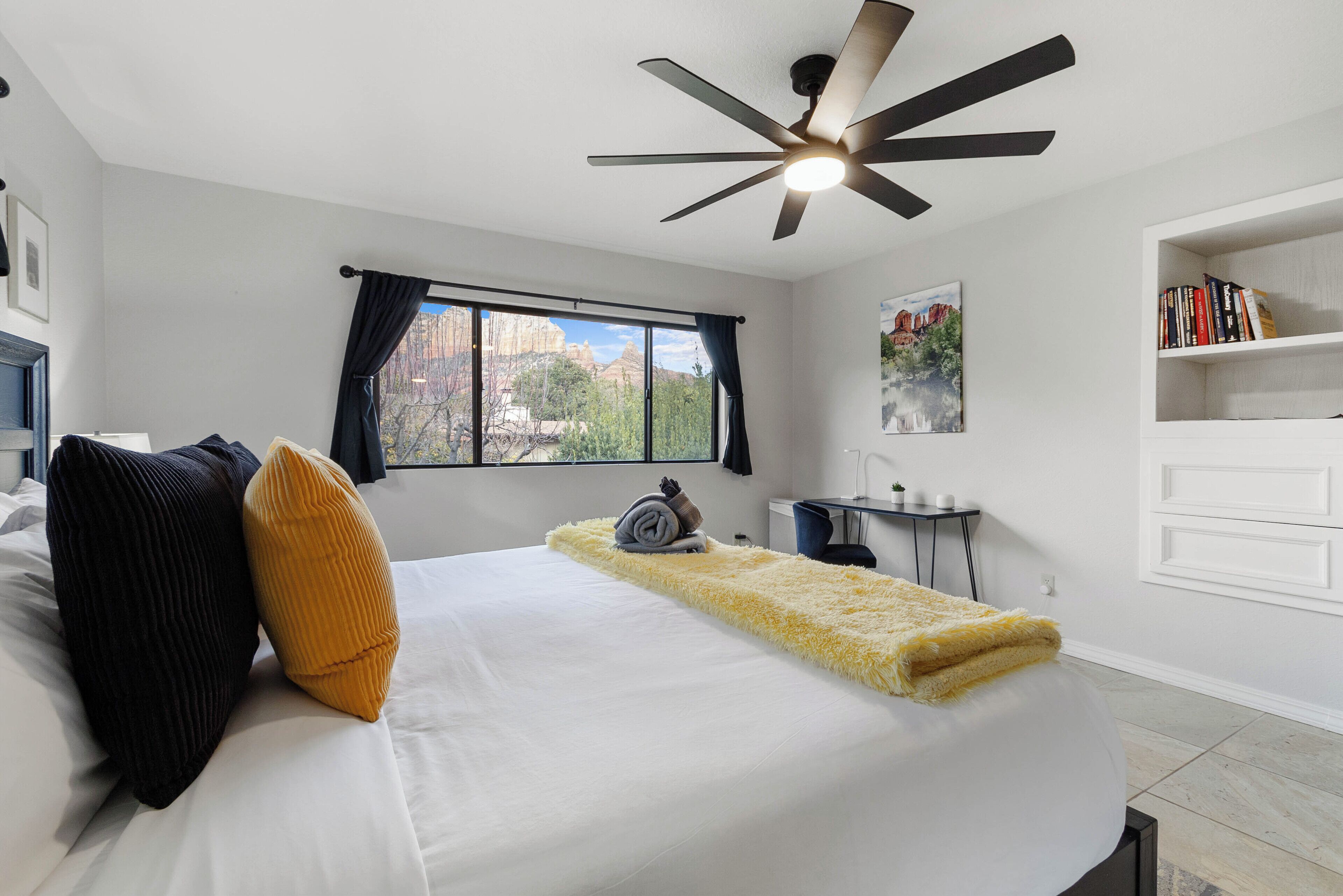 A bedroom with a bed, a desk, a chair, a ceiling fan, and a window with curtains.