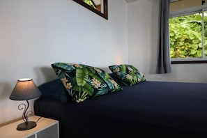 A bedroom with a bed, two pillows with a tropical print, a bedside lamp, and a window with a view of greenery.