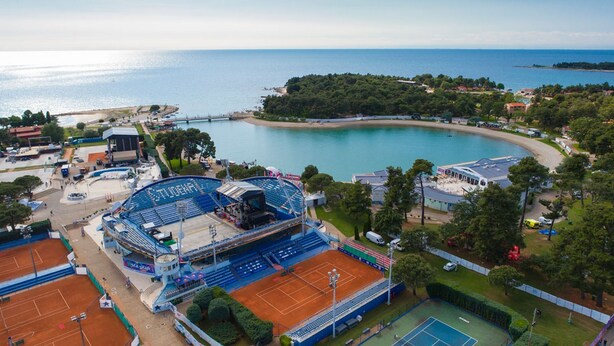 An aerial view of a coastal sports complex with a large stadium, tennis courts, and a beach area.