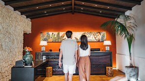 Um casal caminhando em direção a um balcão de recepção em um hall com parede de pedra, teto de madeira e uma grande planta.