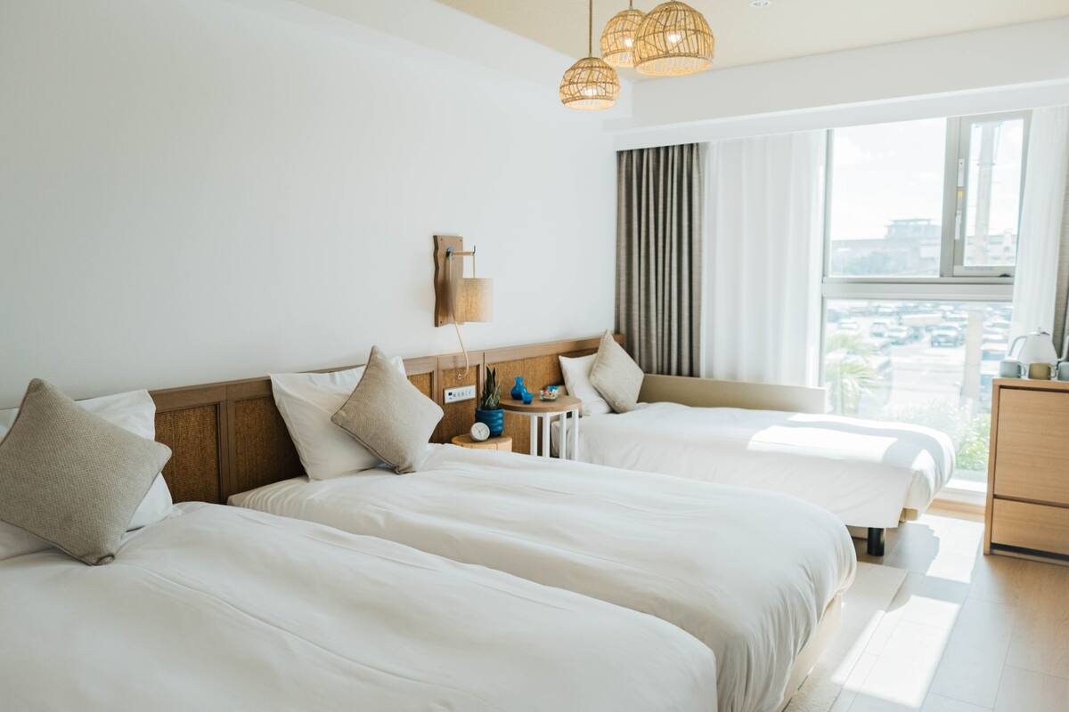 A hotel room with two beds, a small table with a clock, and a view of the city through large windows.