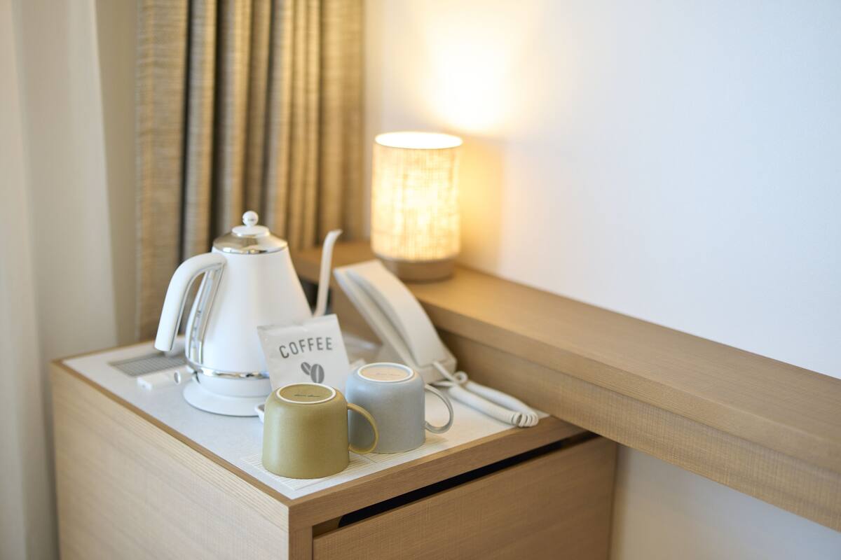 A neatly arranged bedside table with a coffee maker, two mugs, a telephone, and a lit lamp.