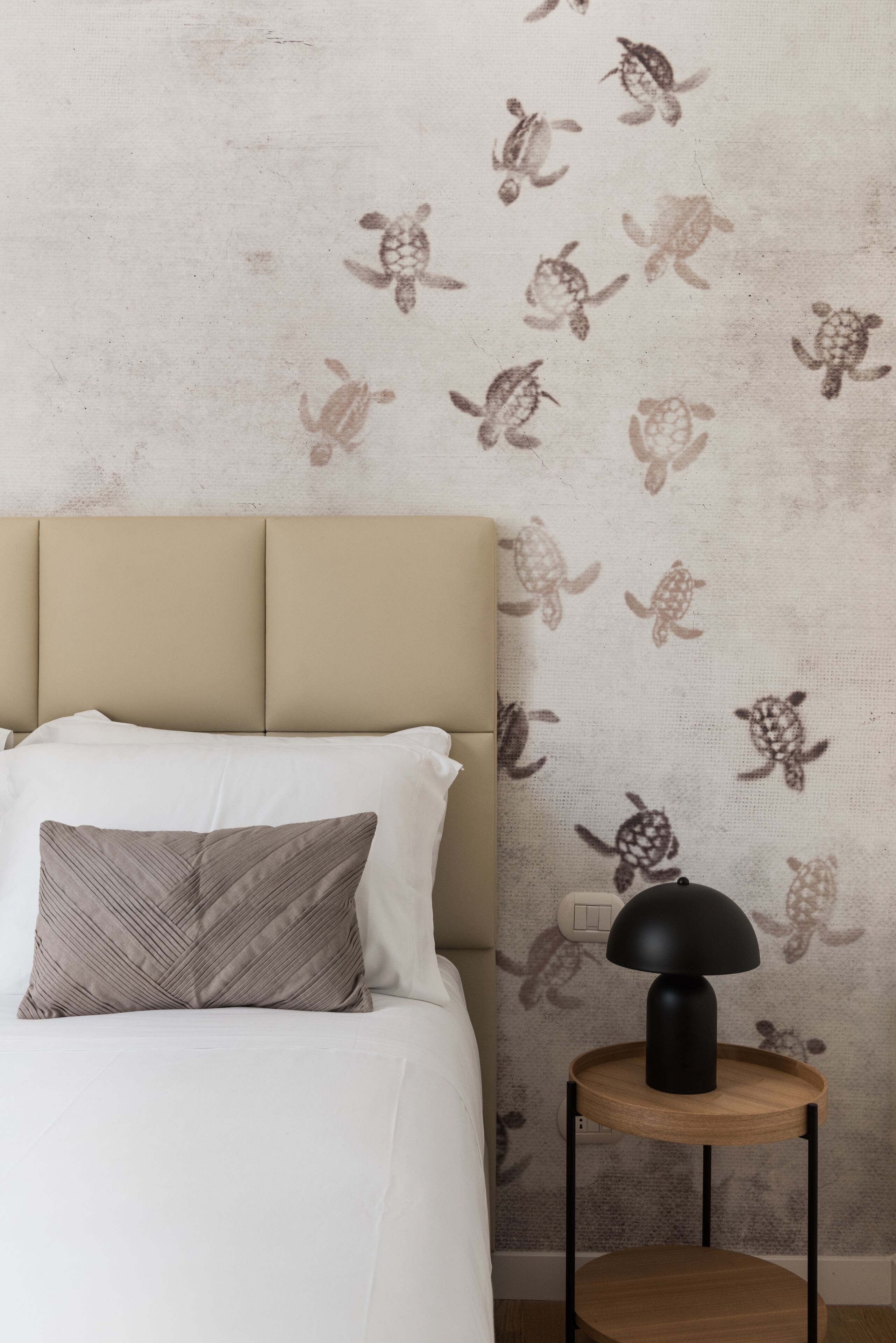 Une chambre à coucher comprenant un lit, des oreillers, une tête de lit, un mur orné d’un motif représentant des tortues, une table de chevet avec une lampe et une prise de courant.