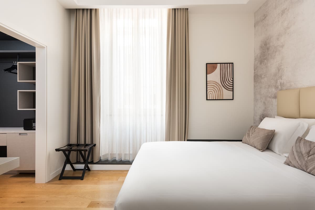 A modern hotel room with a large bed, a built-in wardrobe, and a view of the outdoors through sheer curtains.