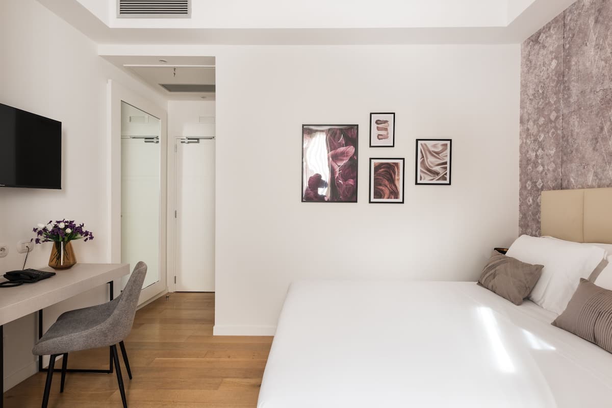 A modern hotel room with a bed, a desk with a chair, and a TV. There are three framed pictures on the wall.