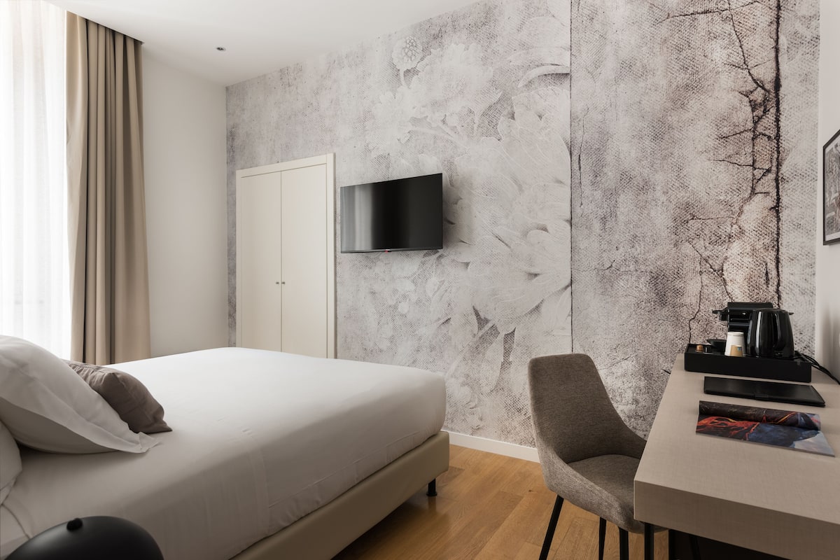 A modern hotel room with a bed, a desk with a chair, a television mounted on the wall, and a door.