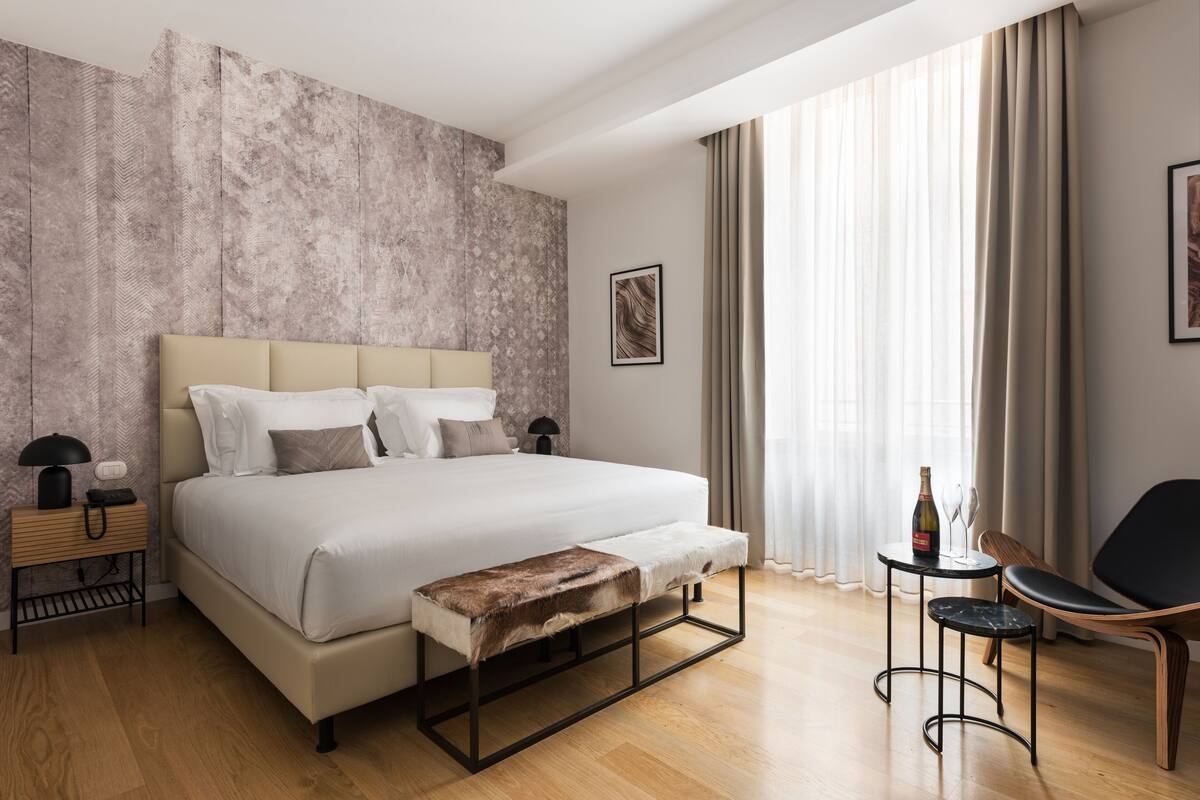 A modern hotel room with a large bed, a bench, a small table with a bottle, and a chair.