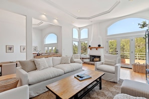 A spacious living room with a large sofa, a coffee table, and a fireplace. There are multiple windows providing natural light and a view of the outdoors. - Santa Barbara Hideaway with Terraced Suites (Santa Barbara)