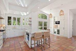 A modern kitchen with white cabinetry, a large island with bar stools, and a view of greenery through the windows. - Santa Barbara Hideaway with Terraced Suites (Santa Barbara)