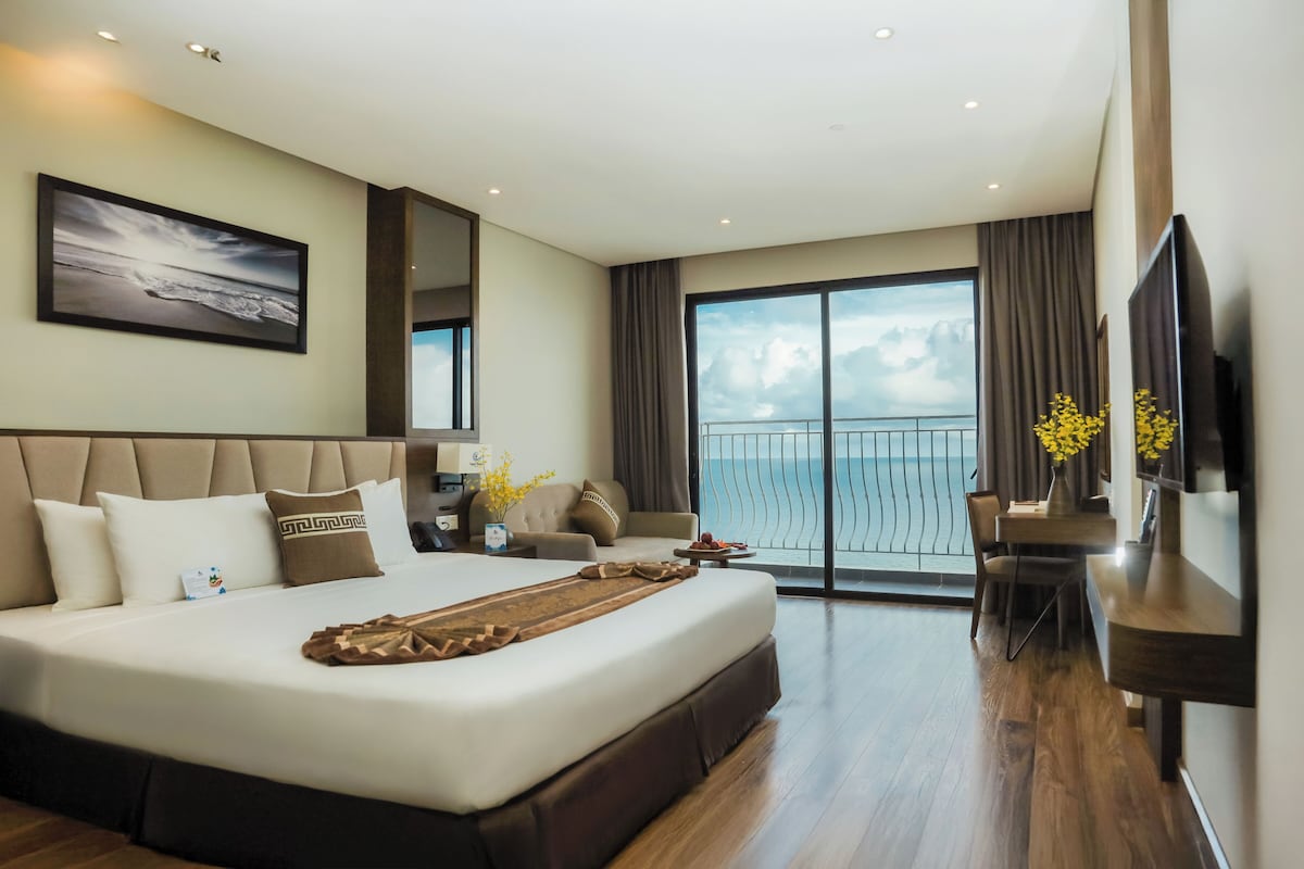 Habitación de hotel moderna con una cama grande, un televisor de pantalla plana, un balcón con vistas al mar y un escritorio con un florero.