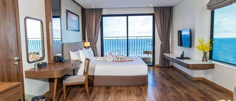 A hotel room with a large bed, a desk, a chair, a TV, and a view of the sea.