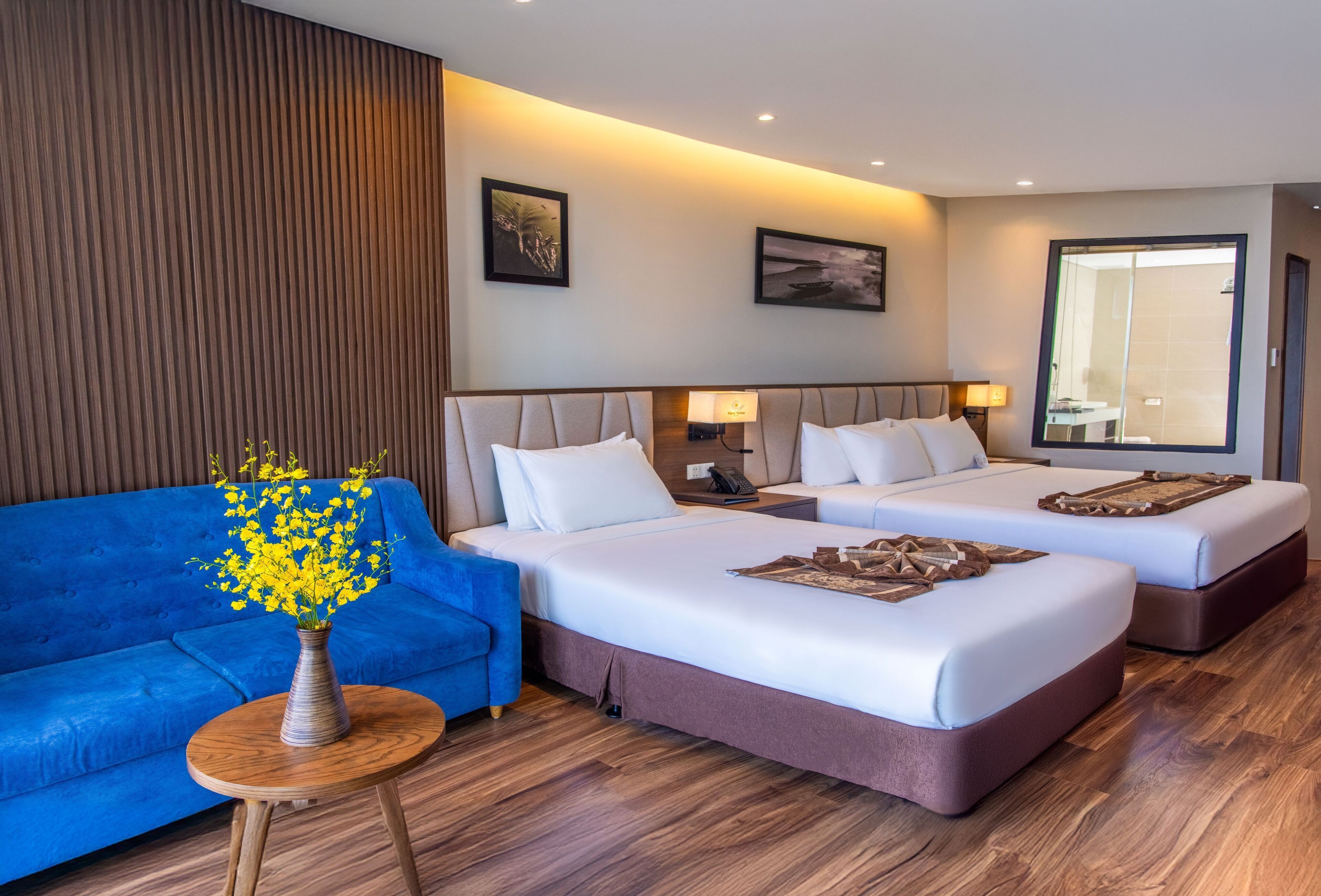 A hotel room with two beds, a blue sofa, a wooden side table with a vase of yellow flowers, and a large mirror.
