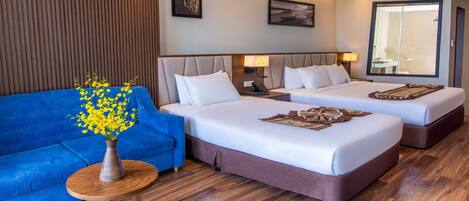 A hotel room with two beds, a blue sofa, a wooden side table with a vase of yellow flowers, and a large mirror.