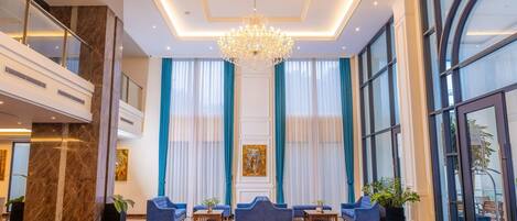 A spacious lobby with a grand chandelier, large windows with blue curtains, and a marble floor.