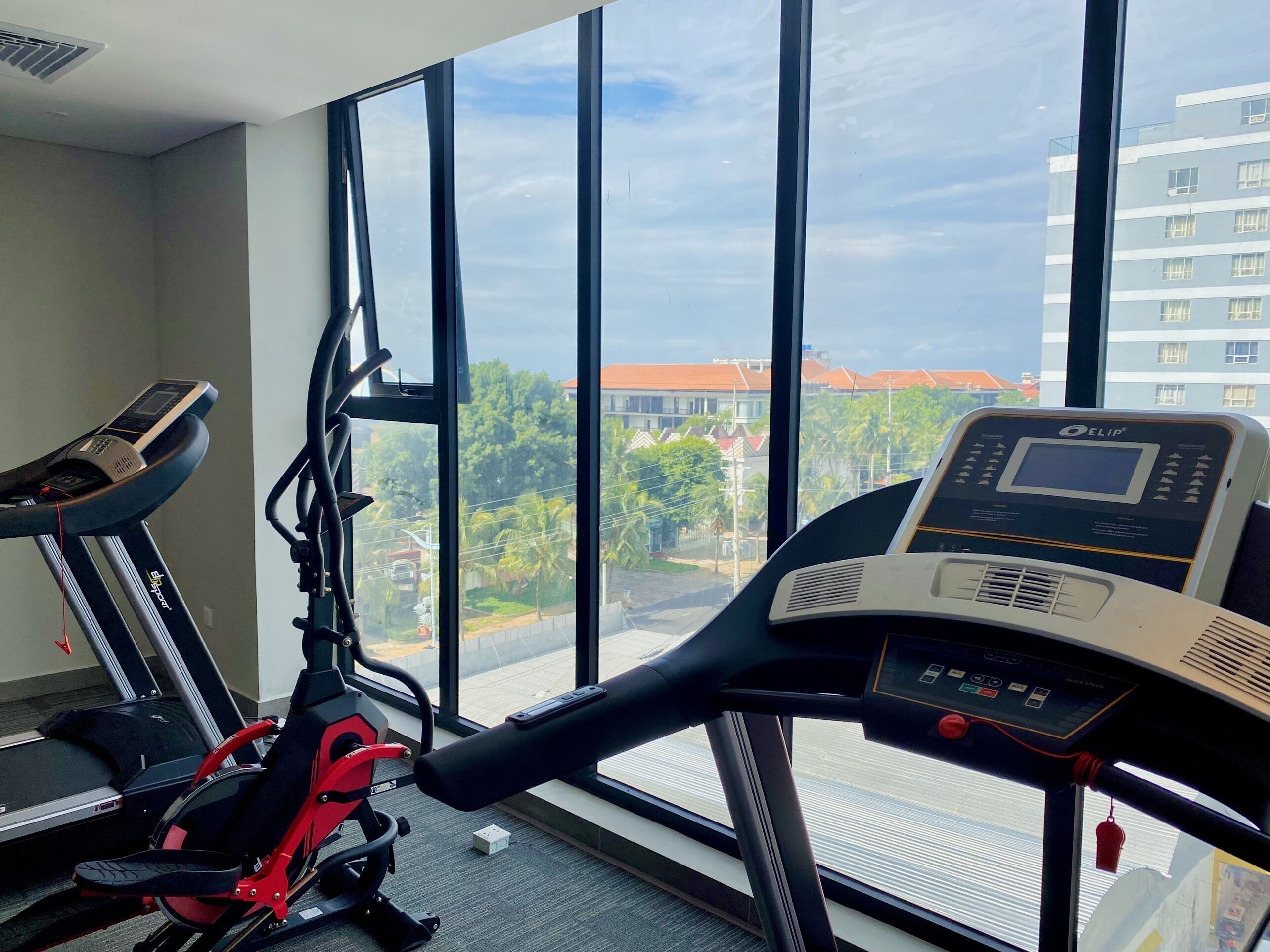 A gym with a treadmill and an elliptical machine, large windows with a view of buildings and trees.