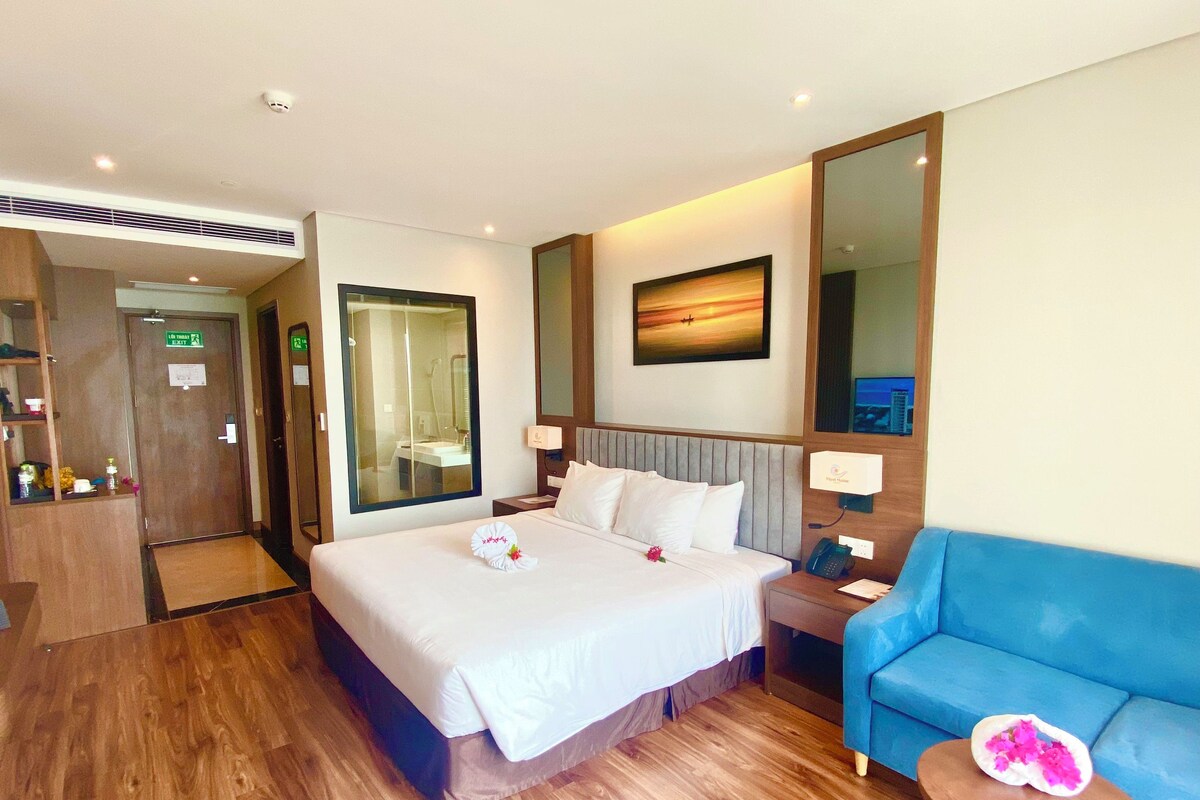 Un cuarto de hotel con una cama grande, un sofá azul, una pequeña mesa con una flor y un cuadro en la pared.
