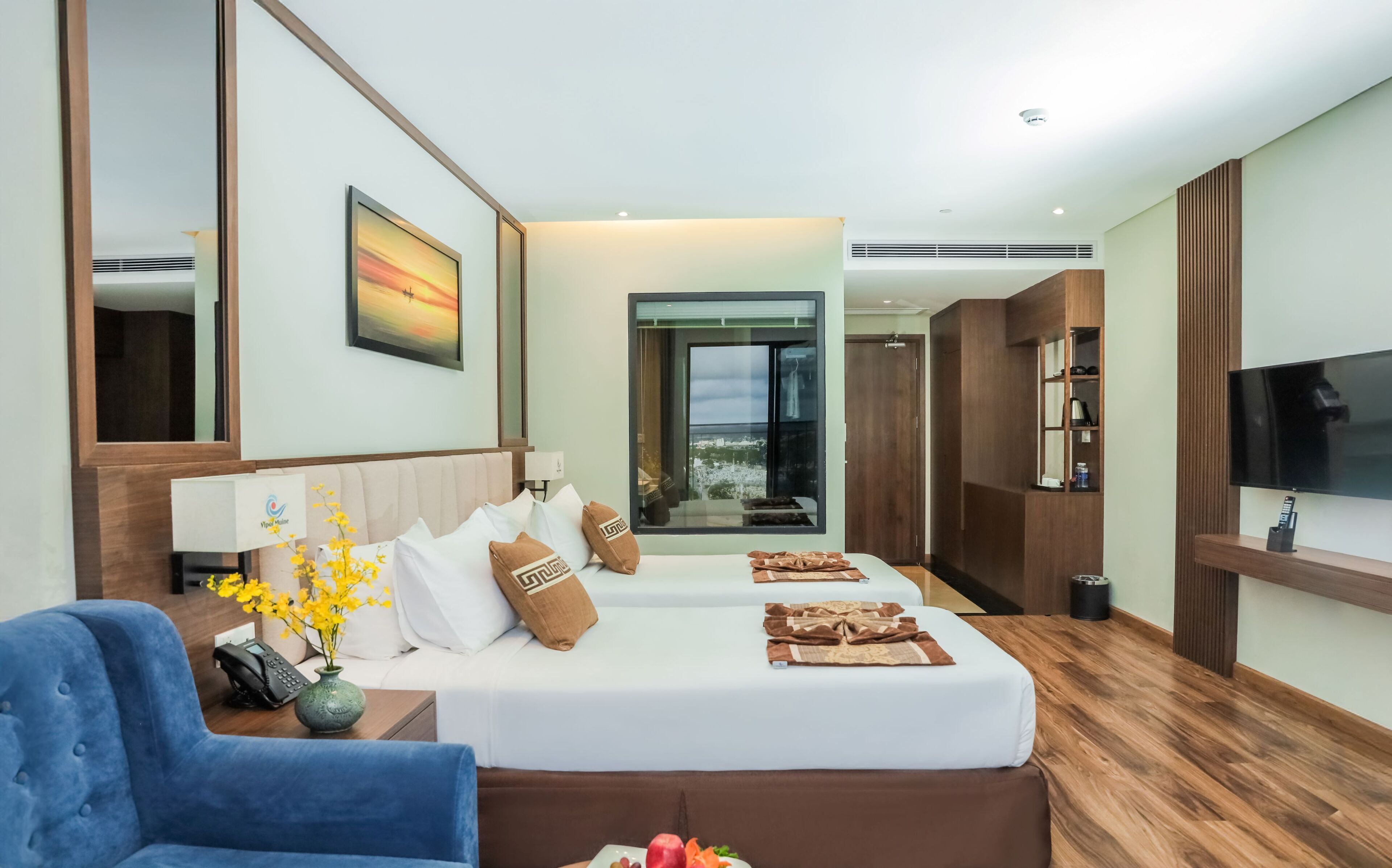 A hotel room with a large bed, a TV, a desk with a phone, and a view of the city through a large window.