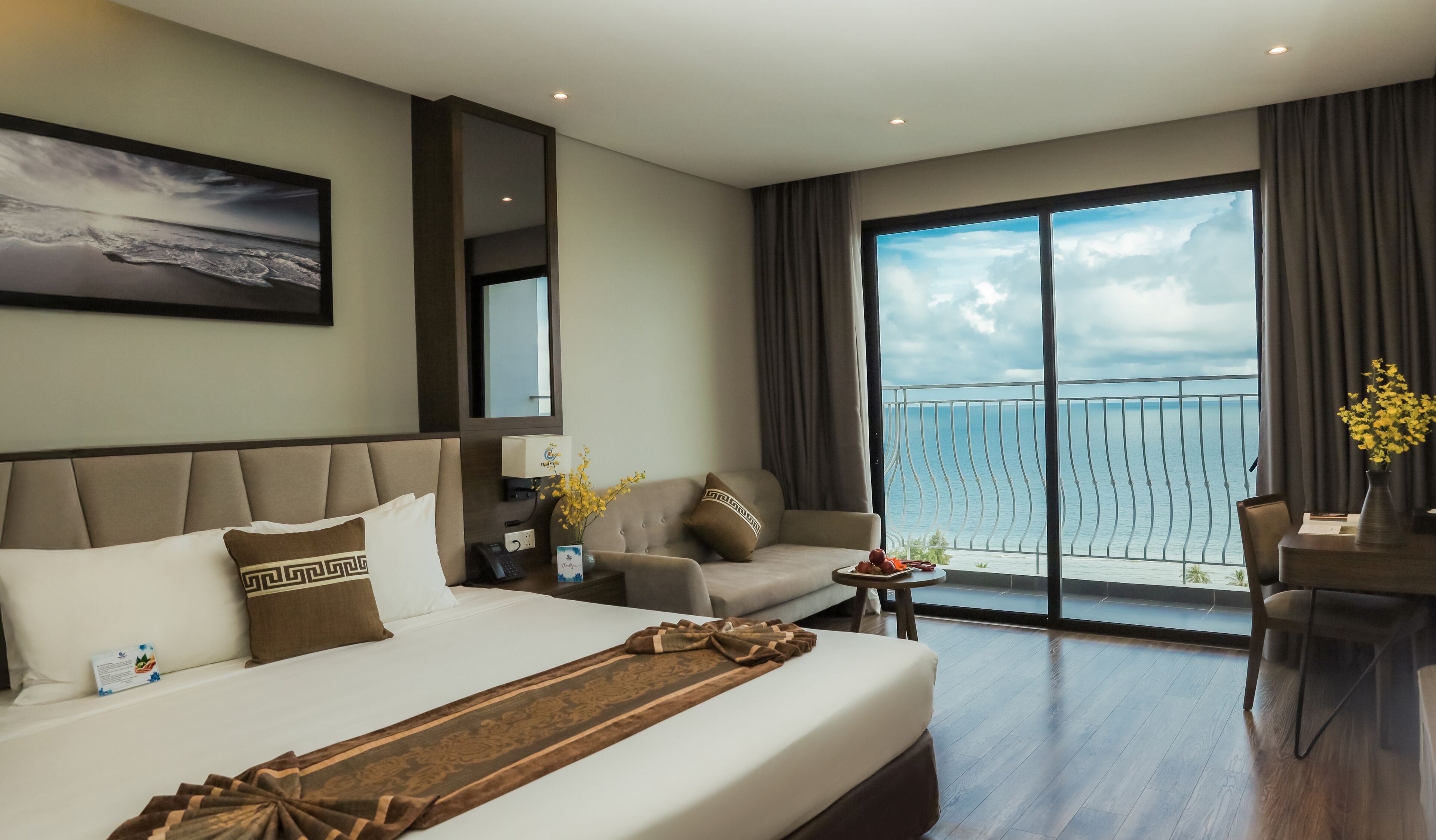 A hotel room with a large bed, a sofa, a dining table, and a balcony with a view of the sea.