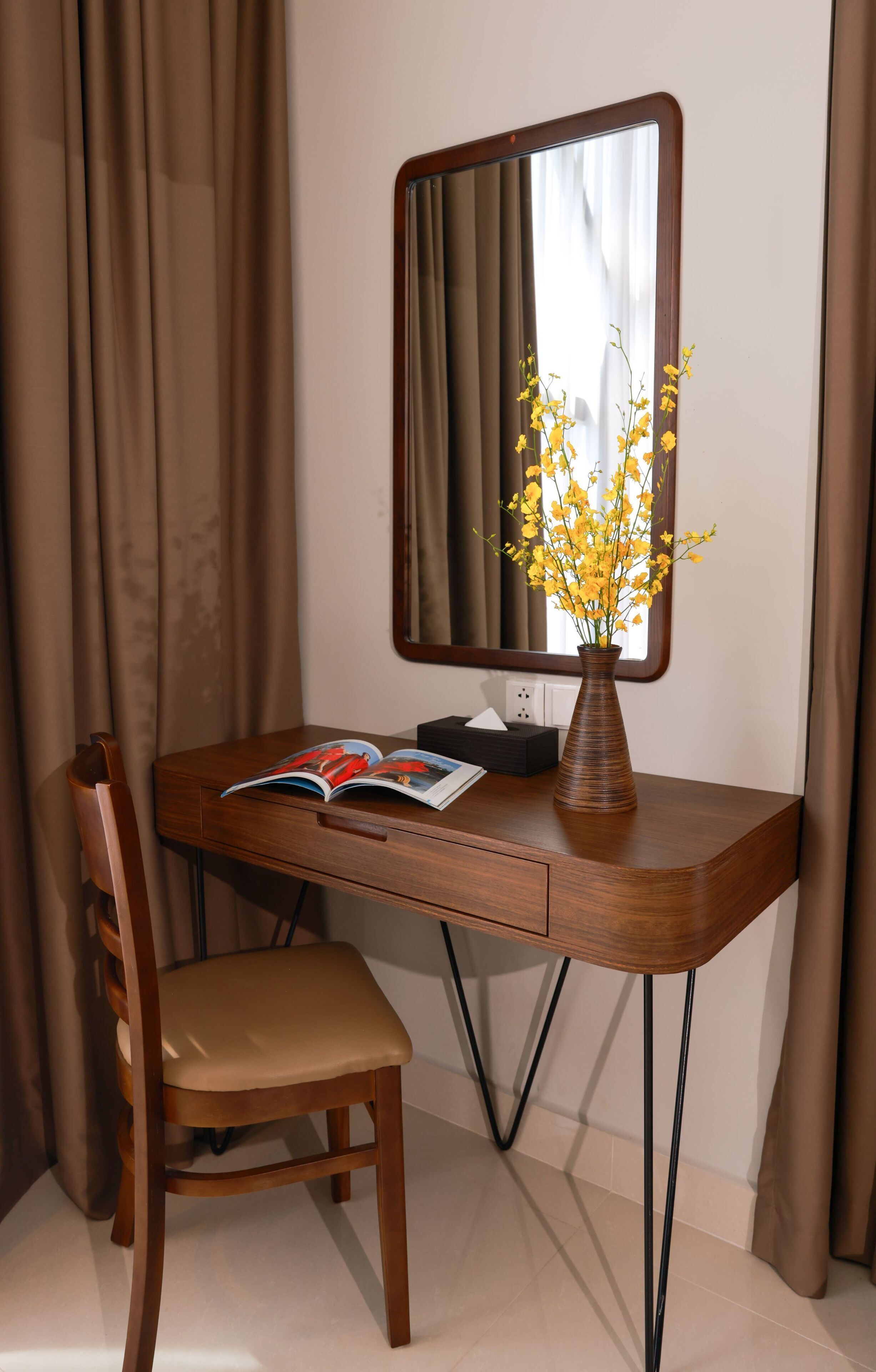 A wooden desk with a mirror, a vase with yellow flowers, and an open magazine.