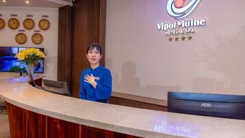 A reception desk with a digital clock, a floral arrangement, and a wall displaying the hotel name and logo.