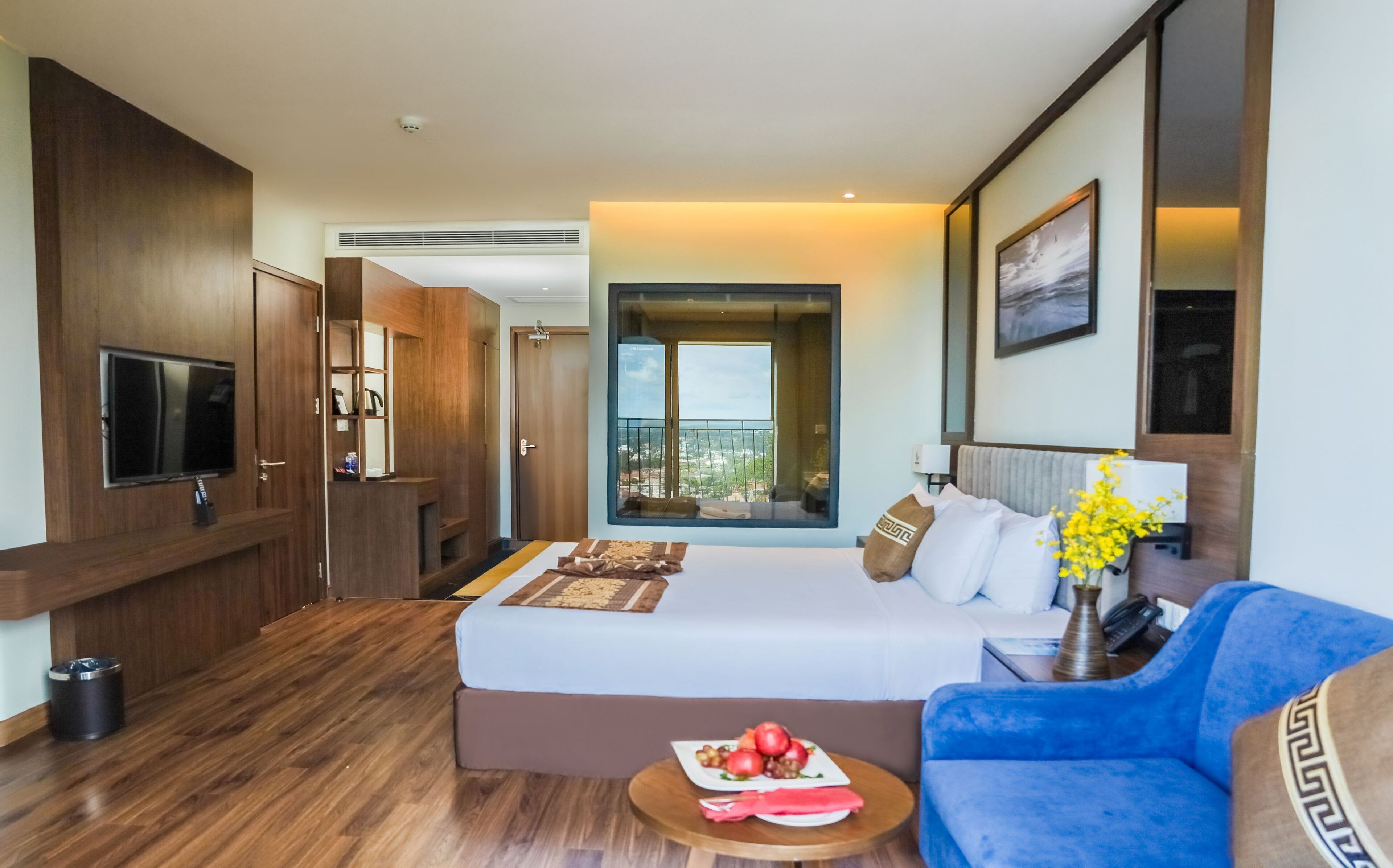 A hotel room with a bed, a TV, a balcony with a city view, and a blue sofa.