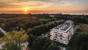 Um edifício moderno com vários andares, rodeado por uma floresta densa, e um pôr do sol ao fundo.