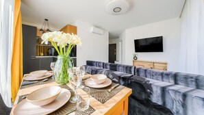 Uma área de refeições com uma mesa posta para quatro pessoas, um sofá, uma TV fixada na parede e uma cozinha ao fundo.