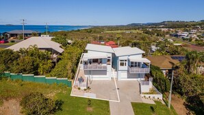 A modern two-story house with a large garage, surrounded by lush greenery and a view of the ocean. - Tea Trees Ocean Views, Pet-Friendly, Walk to shops and Beach (Woolgoolga)