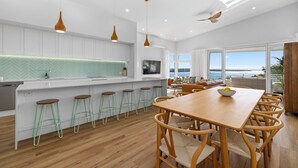 A modern kitchen with white cabinets, a herringbone backsplash, and a dining area with wooden furniture and a view of the ocean. - Tea Trees Ocean Views, Pet-Friendly, Walk to shops and Beach (Woolgoolga)
